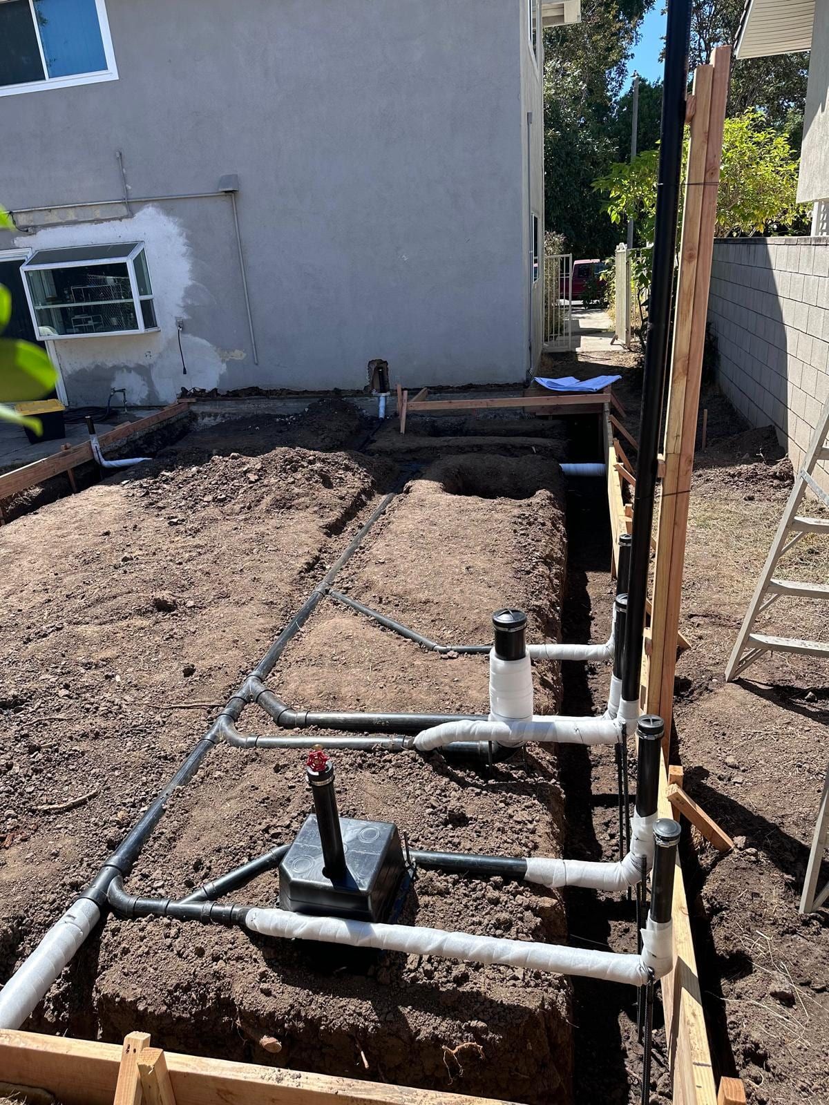 New drain lines with white cleanout risers laid in an open trench beside a house