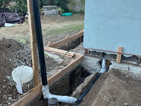 A backyard trench along a house wall with a black drain line, a cleanout and a vent pipe